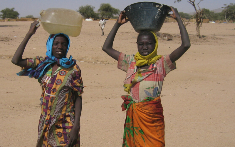 Two adolescent girls in a refugee camp hold buckets of water.